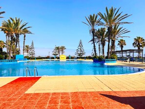 A large outdoor swimming pool with palm trees and a clear blue sky. - Modern, Bright Apartment Right by the Beach (El Mansouria)