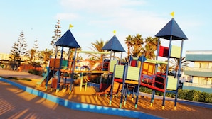 Ein Spielplatz mit verschiedenen Spielgeräten, darunter Rutschen und Kletterbereiche, umgeben von Palmen und blauem Himmel.