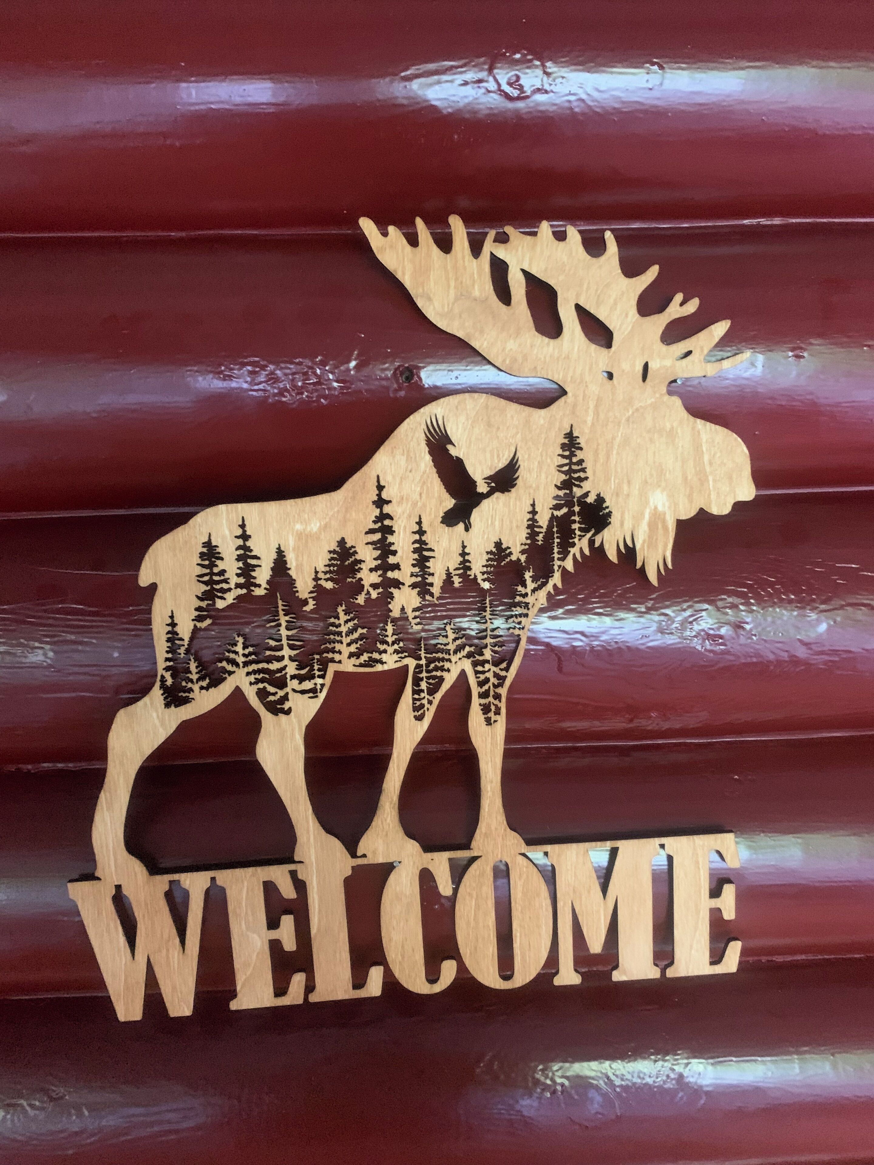 A wooden welcome sign featuring a moose silhouette and forest trees, mounted on a red wooden background.