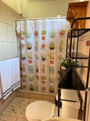 Une salle de bain avec un rideau de douche orné d’un motif de cactus, des toilettes blanches, un porte-serviettes avec une serviette à rayures et une étagère métallique noire sur laquelle est posée une plante.