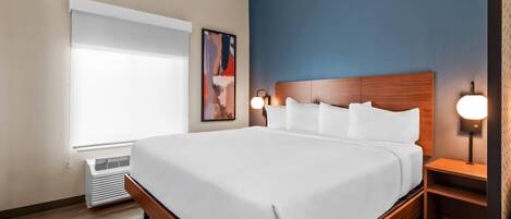 A hotel room with a large bed, a window with blinds, a wall-mounted artwork, and a bedside table with a lamp.