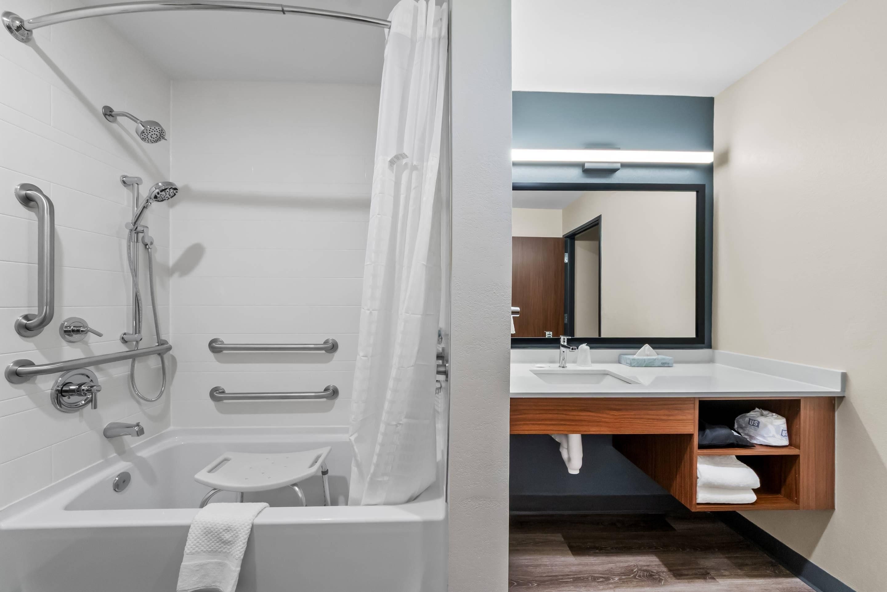 A modern bathroom with a shower, grab bars, a small stool, and a vanity with a mirror and sink.