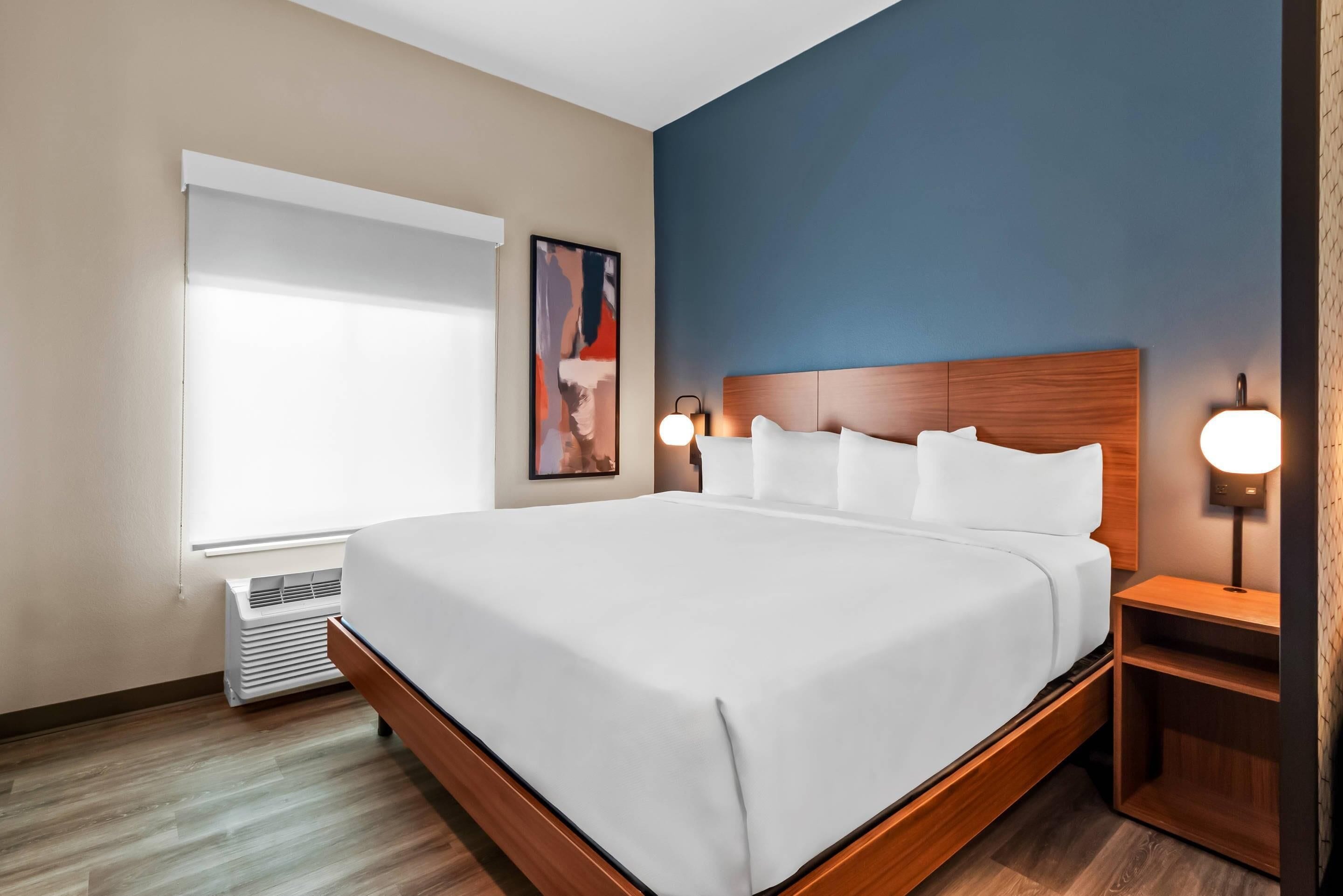 A hotel room with a large bed, a window with blinds, a wall-mounted artwork, and a bedside table with a lamp.