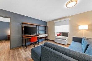 A modern living room with a grey sofa, a wooden desk with a TV, a red office chair, and a window with blinds. - Everhome Suites San Antonio Lackland near SeaWorld by Choice Hotels (San Antonio)