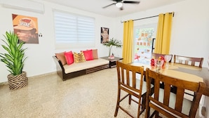Un salon comprenant un canapé, une table à manger avec des chaises, un ventilateur de plafond et une fenêtre avec des stores.