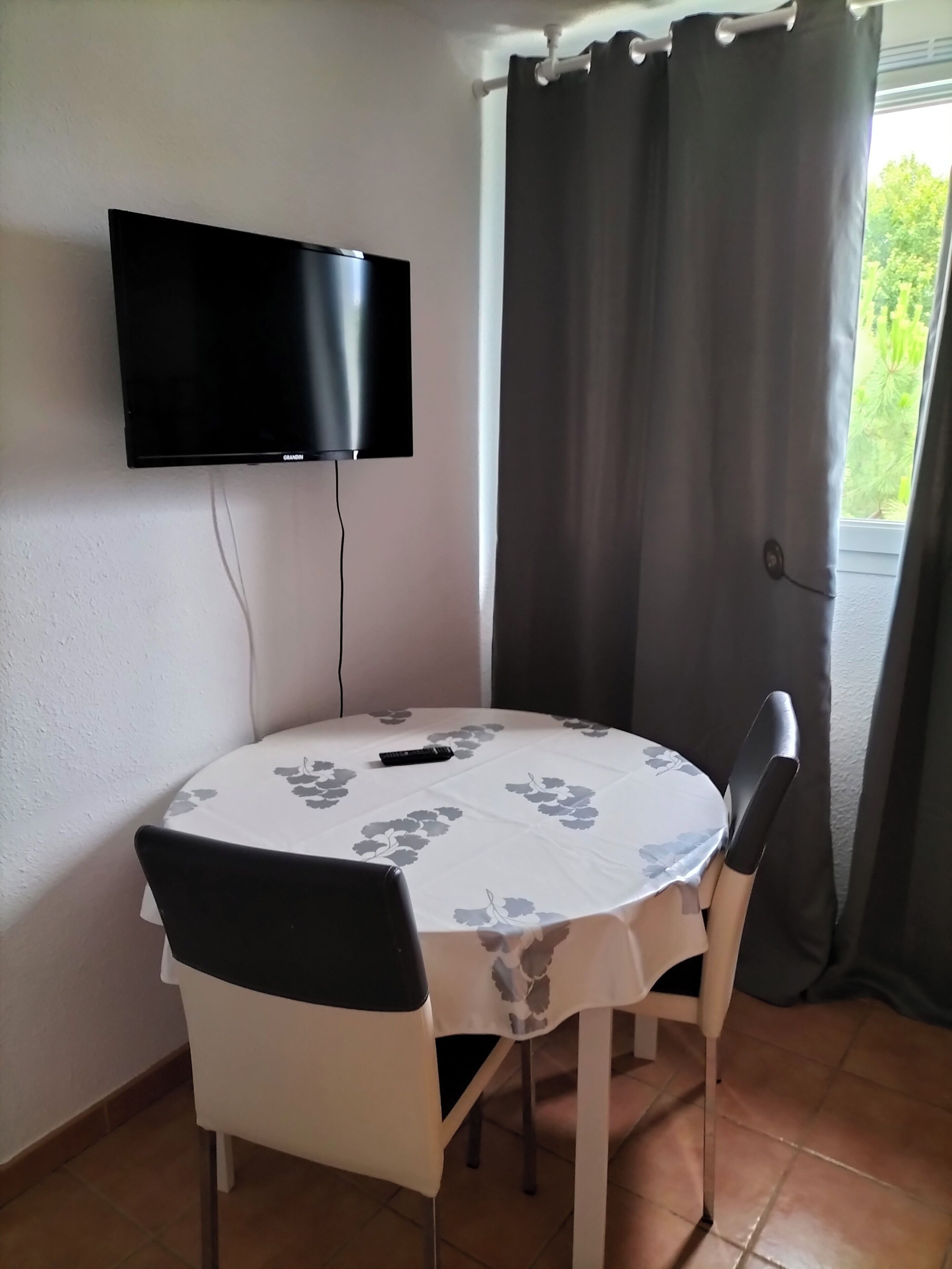 A dining area with a round table covered by a white tablecloth with grey floral patterns, two chairs, a flat-screen TV mounted on the wall, and a window with curtains.