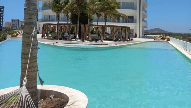 Ein mehrstöckiges Hotel mit Poolbereich, Palmen und blauem Himmel.