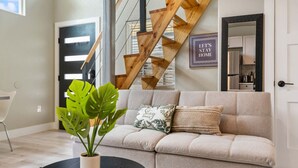 Un salon moderne avec un canapé beige, une plante en pot et une table basse noire. Un escalier en bois avec une rampe noire est visible à l’arrière-plan.