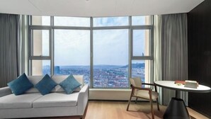A modern hotel room with a large window offering a city view, a sofa with blue cushions, a round table, and a black floor lamp. - Guangsha Hotel (Shengzhou)