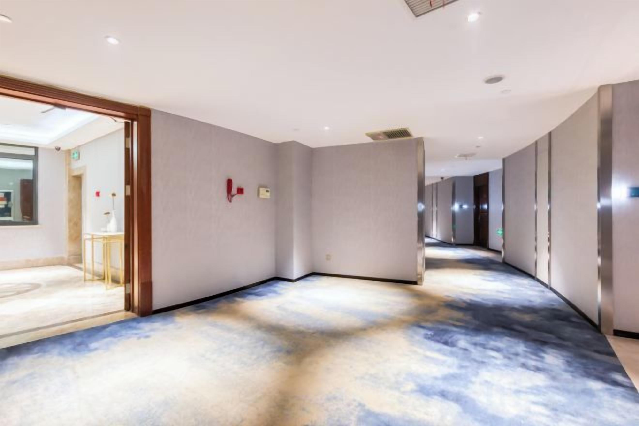 A hotel corridor with a carpeted floor, a fire alarm, and a door leading to a room.