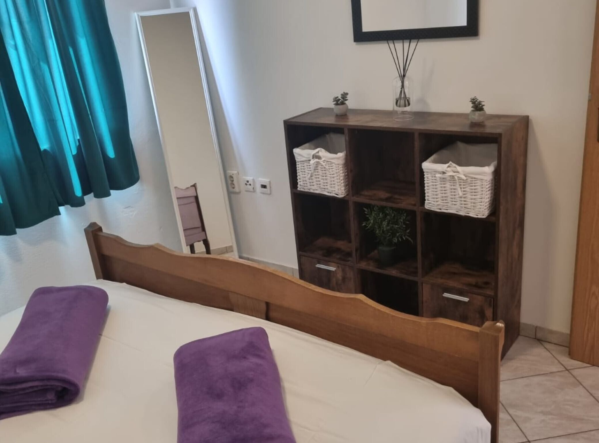 A bedroom with a wooden bed, two purple towels, a wooden nightstand, a mirror, and a wooden wardrobe with baskets.
