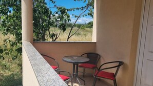 A small outdoor balcony with a table and two chairs, a door to the right, and a view of trees and grass.