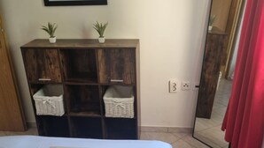 A bedroom with a bed, a wooden wardrobe with baskets, two potted plants, and a mirror with red curtains.