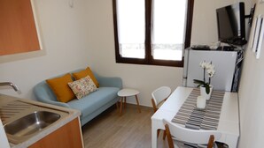 A compact kitchen with a sink, a small dining table with chairs, a sofa with cushions, and a television mounted on the wall.