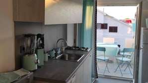 A compact kitchen with a sink, a kettle, and a stove. There is a window with a curtain, and a balcony with chairs.