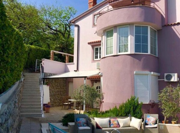 A two-story house with a stone staircase leading to a balcony, outdoor seating area, and a covered patio.