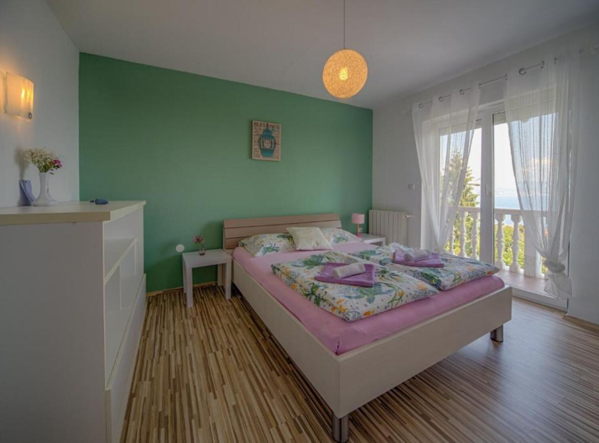 Quarto com cama, mesa de cabeceira, luminária na parede, janela com cortinas e vista para o verde lá fora.