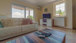 Un salón con un sofá, una mesa de centro con mandos a distancia, un televisor sobre un mueble bajo y ventanas grandes con vistas al cielo.
