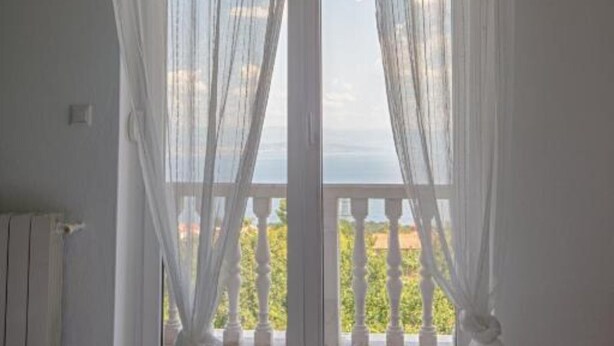 Una habitación con un ventanal grande, cortinas blancas traslúcidas, vistas a follaje y un paisaje lejano.