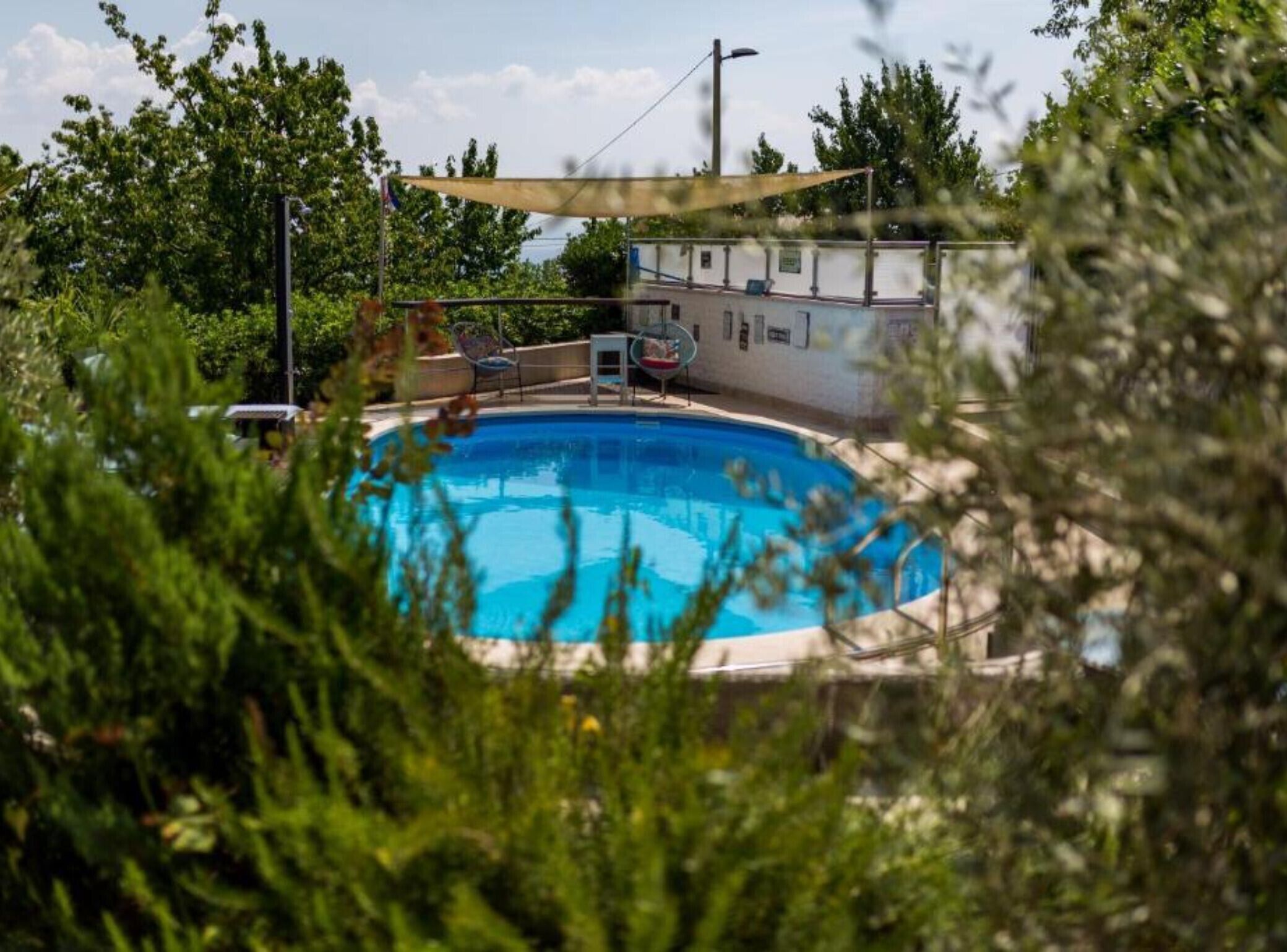 A swimming pool surrounded by greenery, with a covered seating area and a building in the background.