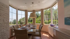 Un comedor con una mesa grande dispuesta para cuatro personas, rodeada de sillas, ventanas amplias que ofrecen vistas a zonas verdes, y una lámpara colgante sobre la mesa.