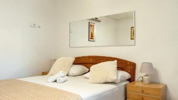 A neatly made bed with a quilt, pillows, and rolled towels. There is a wooden nightstand with a lamp and a small decorative item. The room has a simple, clean design with a tiled floor.