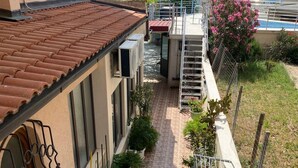 Un edificio residencial con techo de tejas, un pequeño jardín con plantas en macetas, una escalera que conduce a un balcón y vista al mar.