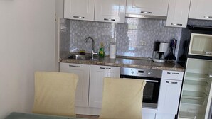 Cocina compacta con gabinetes blancos, protector contra salpicaduras de azulejos, un lavamanos, cafetera y microondas. Hay dos sillas en una mesa.
