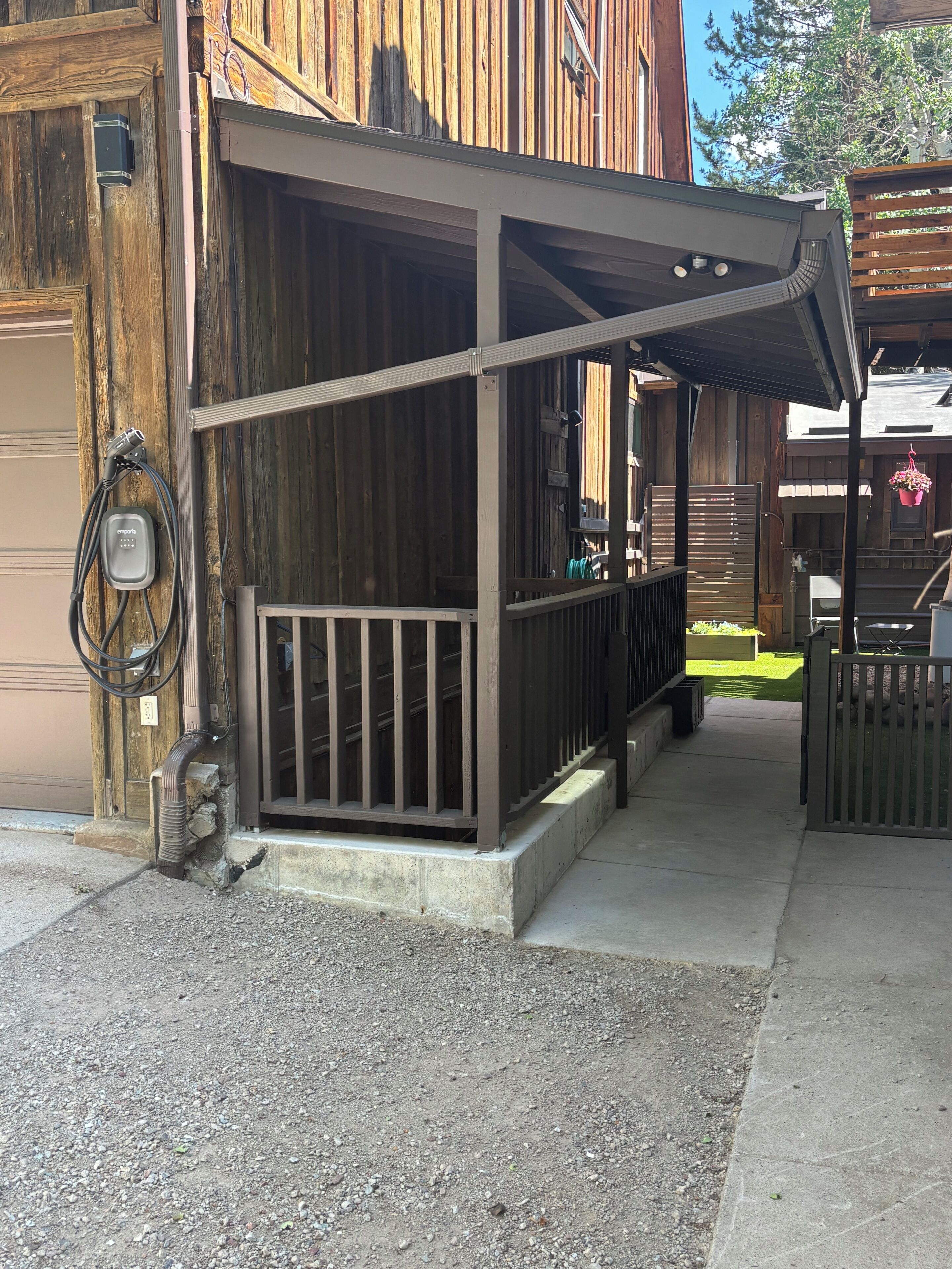 Un espacio exterior techado con una terraza de madera, un sendero de hormigón y una verja metálica.