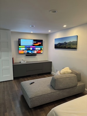 Una sala de estar moderna con un televisor de pantalla plana montado en la pared, un sofá gris, una puerta blanca y un cuadro de montañas.