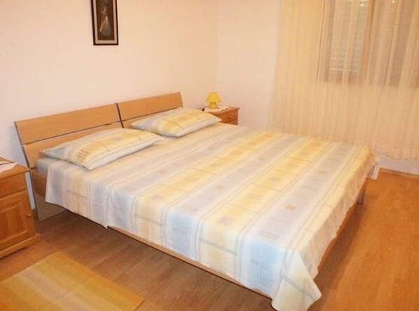 A single bed with a wooden headboard, a nightstand with a lamp, and a window with sheer curtains.