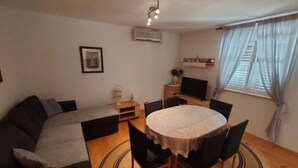 Ein Wohnzimmer mit einer Couch, einem runden Tisch mit Stühlen, einem Fernseher, einem kleinen Beistelltisch und einem Fenster mit Vorhängen.