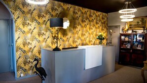A reception area with a patterned wallpaper, a reception desk, a standing lamp, a decorative bird sculpture, and a bookshelf with books and decorative items. - Hotel Castel Moor (Le Croisic)