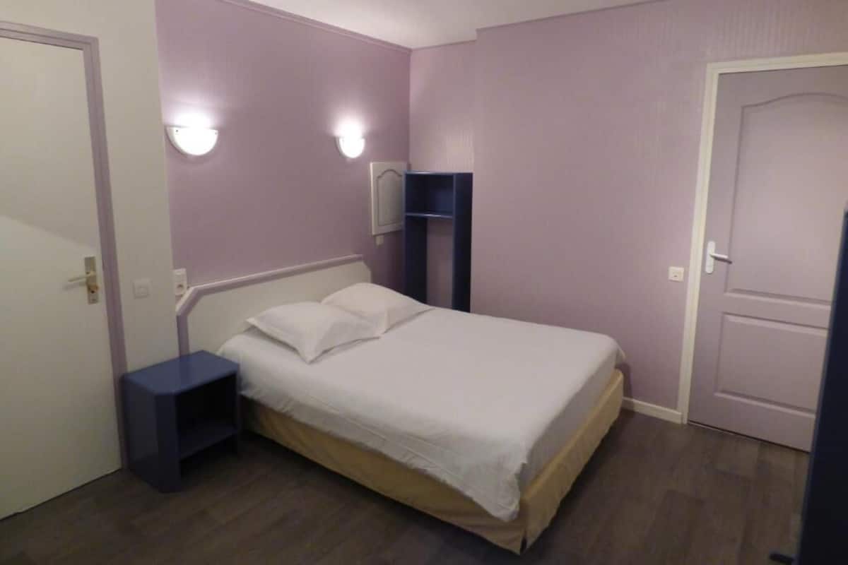 A bedroom with a single bed, a nightstand, a wall-mounted shelf, and a door.