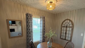 A dining room with a round wooden table, four wooden chairs, a vase with flowers, patterned curtains, a mirror, and a decorative ceiling light. - The Gatehouse, 2-bedroom house in charming Gamaliel with WiFi, AC, pet friendly. (Gamaliel)