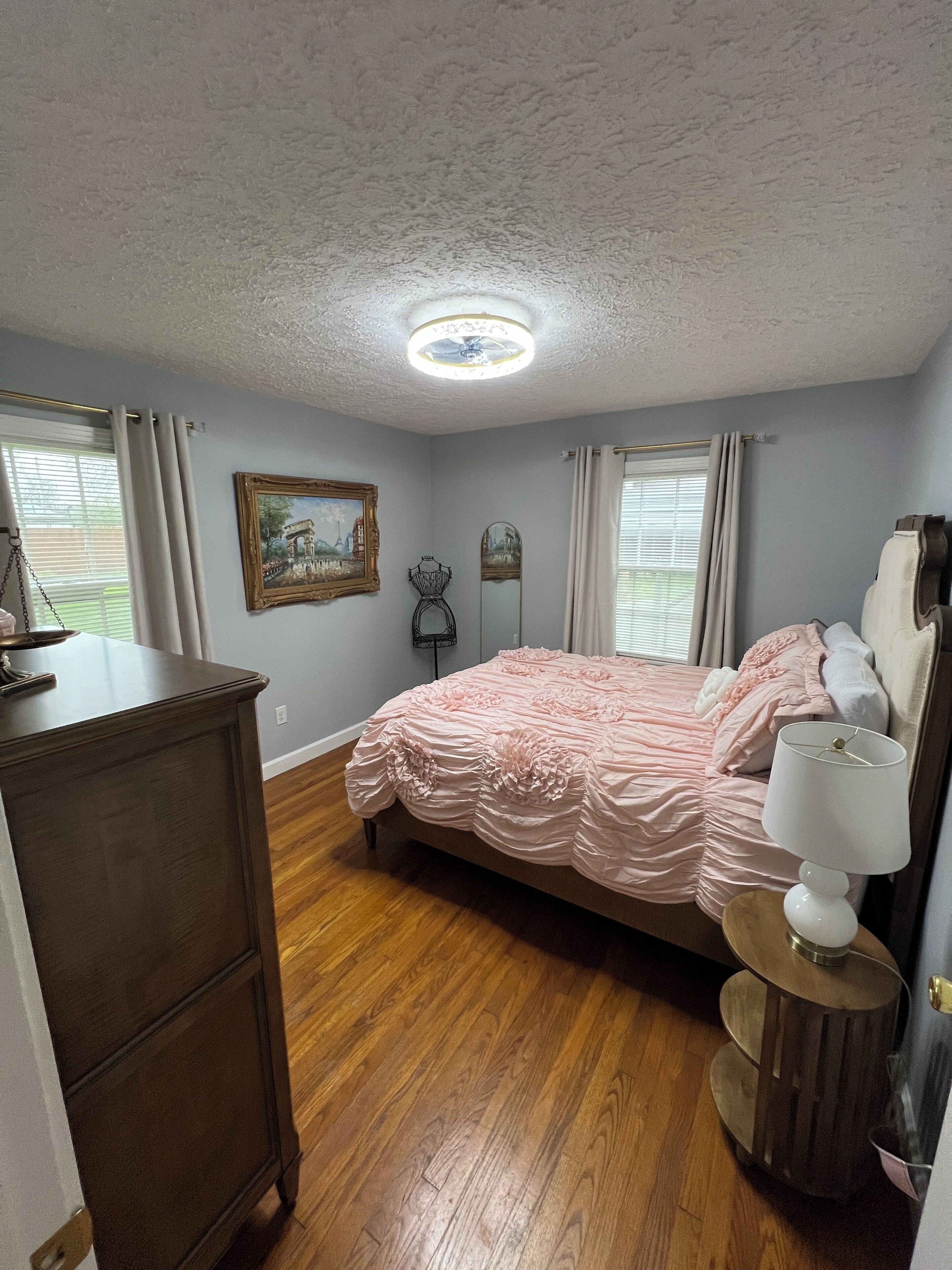 A bedroom with a wooden floor, a bed with pink bedding, a dresser, a bedside lamp, and a painting on the wall.