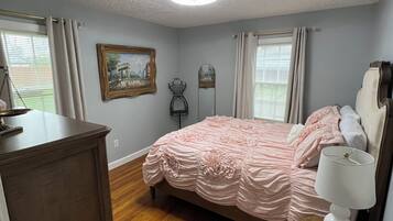 A bedroom with a wooden floor, a bed with pink bedding, a dresser, a bedside lamp, and a painting on the wall.