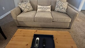 A living room with a beige sofa, patterned cushions, a wooden coffee table with a remote control, and a chandelier. - The Gatehouse, 2-bedroom house in charming Gamaliel with WiFi, AC, pet friendly. (Gamaliel)