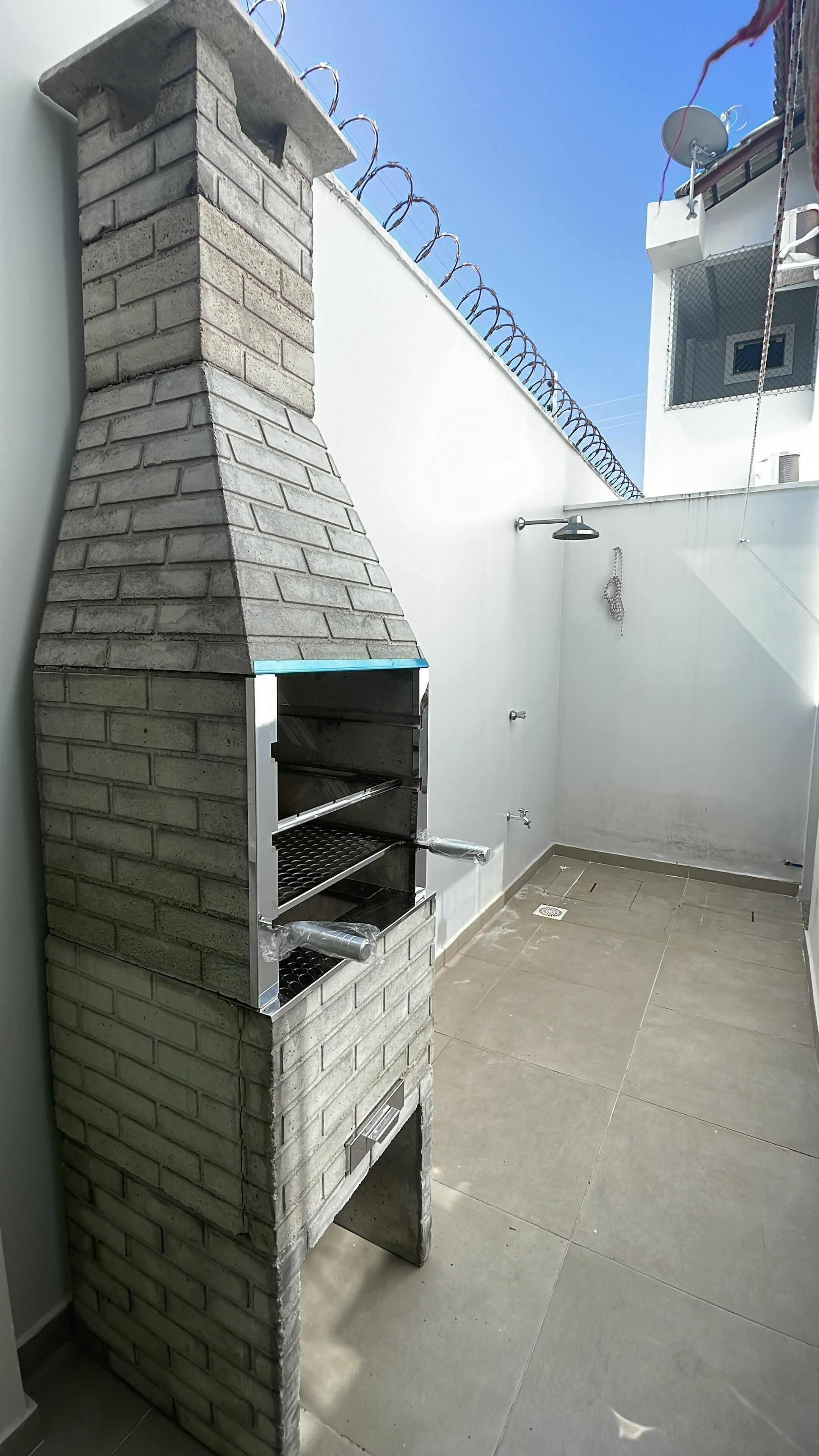 A brick-built outdoor oven with a chimney, a white wall with a barbed wire fence, and a satellite dish. - Cozy house facing east, in a gated community - Salinópolis/PA (Salinópolis)
