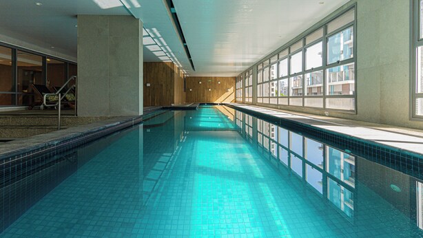 A long indoor swimming pool with clear blue water, large windows on one side, and a staircase on the other.