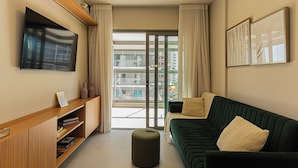 A modern living room with a green sofa, a flat-screen TV, a wooden console table with books, and a sliding glass door leading to a balcony.