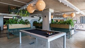 A pool table in a modern indoor space with hanging wicker lamps, green seating, and potted plants.