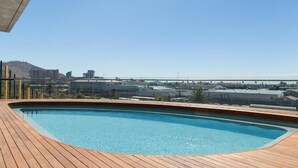 A rooftop pool with a wooden deck and a cityscape view in the background.