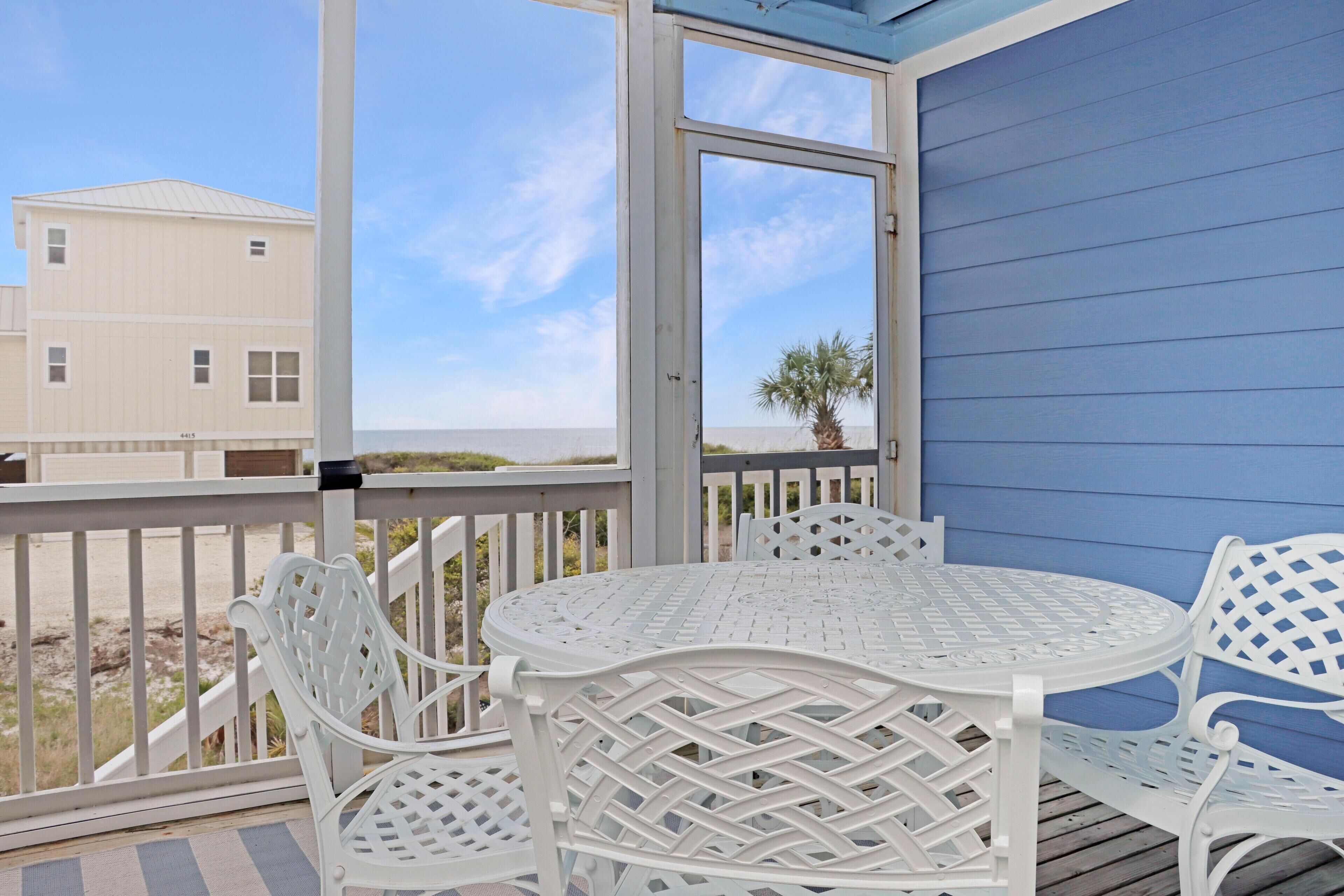 A covered outdoor area with a round table and white chairs, overlooking a beach and a building.