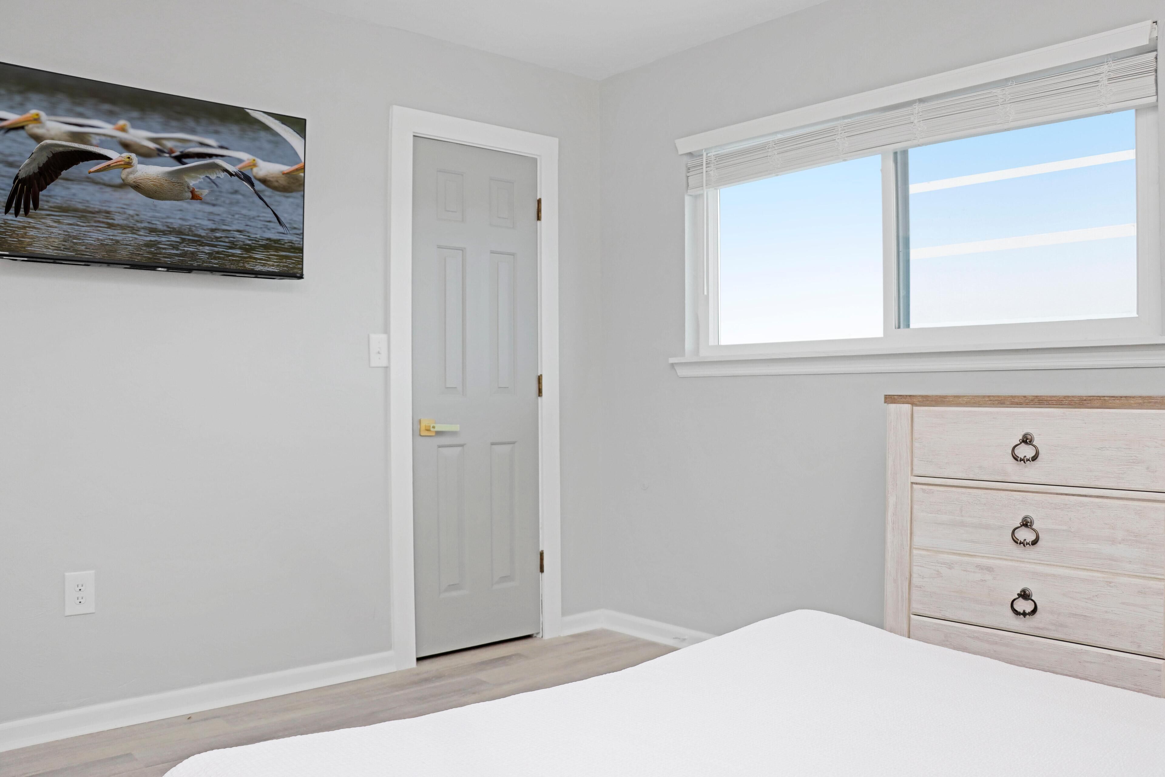 A bedroom with a bed, a dresser, a door, a window, and a wall-mounted picture of pelicans.