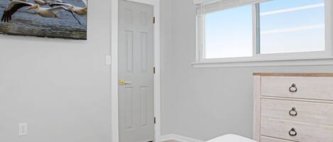 A bedroom with a bed, a dresser, a door, a window, and a wall-mounted picture of pelicans.