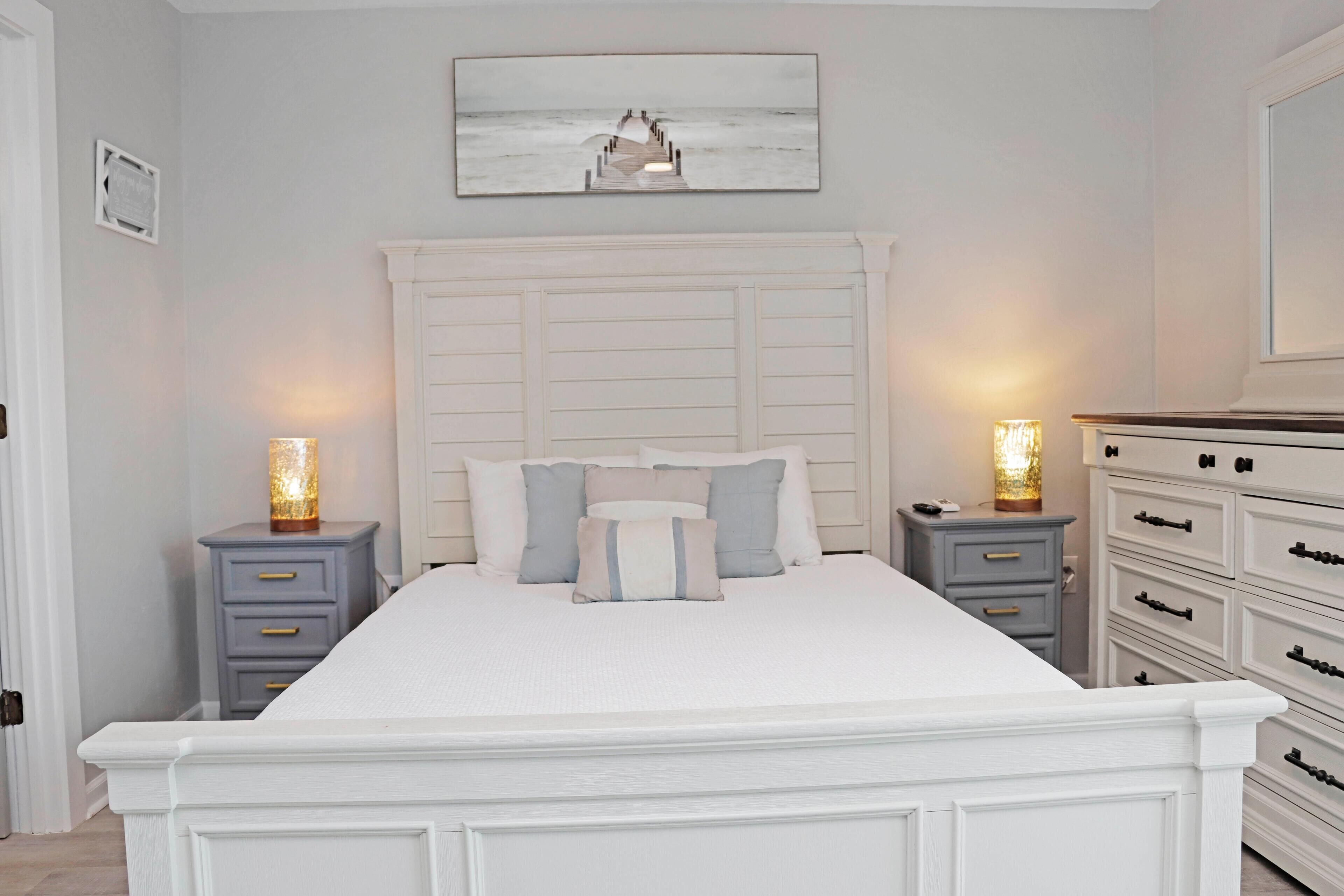 Une chambre à coucher avec un grand lit, deux tables de chevet avec des lampes, une commode et un tableau encadré au mur.