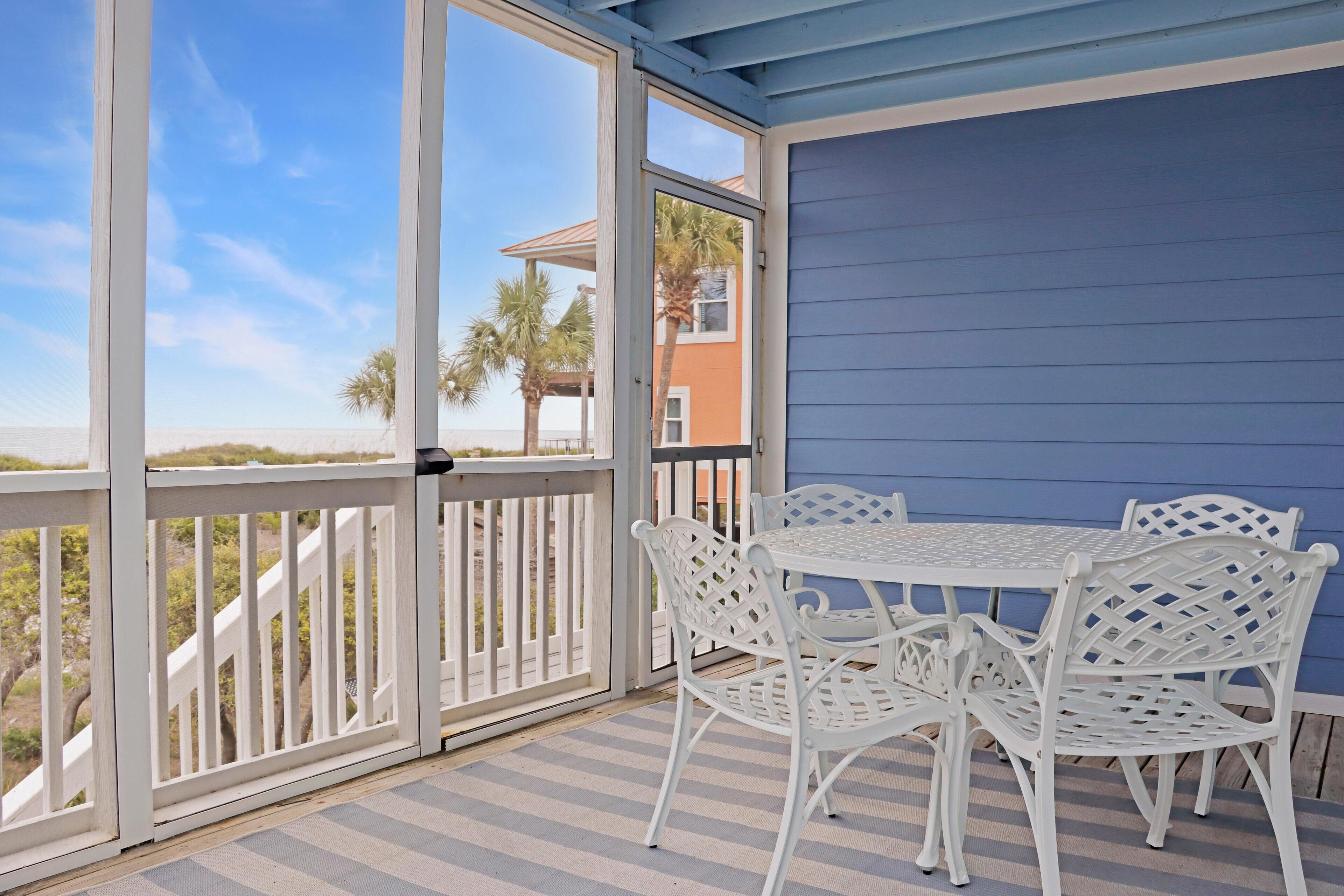 Un espace extérieur abrité, avec une table et des chaises blanches, une vue sur la plage et un bâtiment à la façade bleue.
