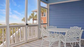 Un espace extérieur abrité, avec une table et des chaises blanches, une vue sur la plage et un bâtiment à la façade bleue.