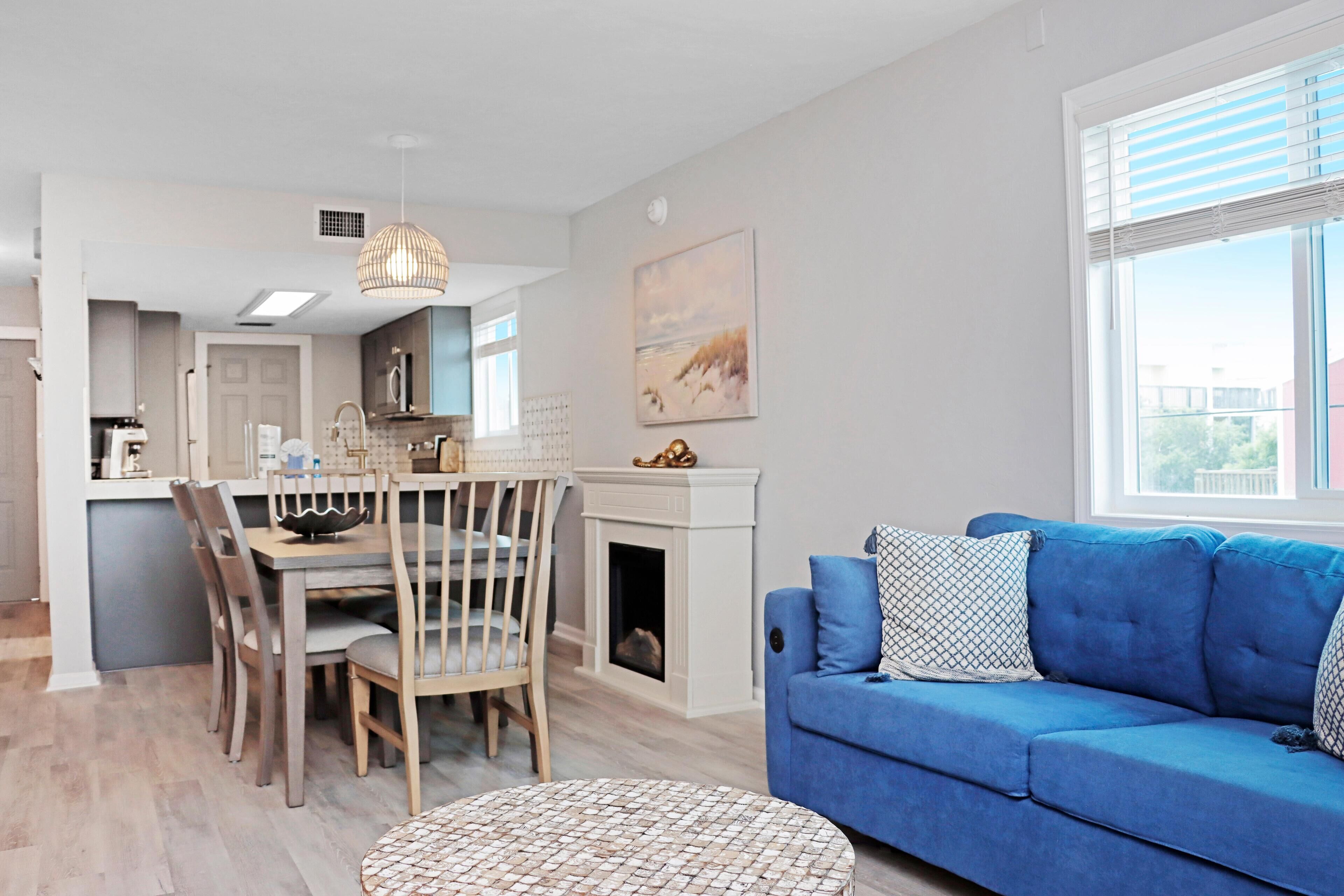 A modern living room with a blue sofa, a dining table with chairs, a fireplace, and a large window with blinds.